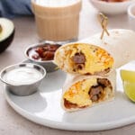 A hearty breakfast burrito filled with scrambled eggs, sausage, cheese, and hash browns, served with lime wedges, salsa, and sour cream.